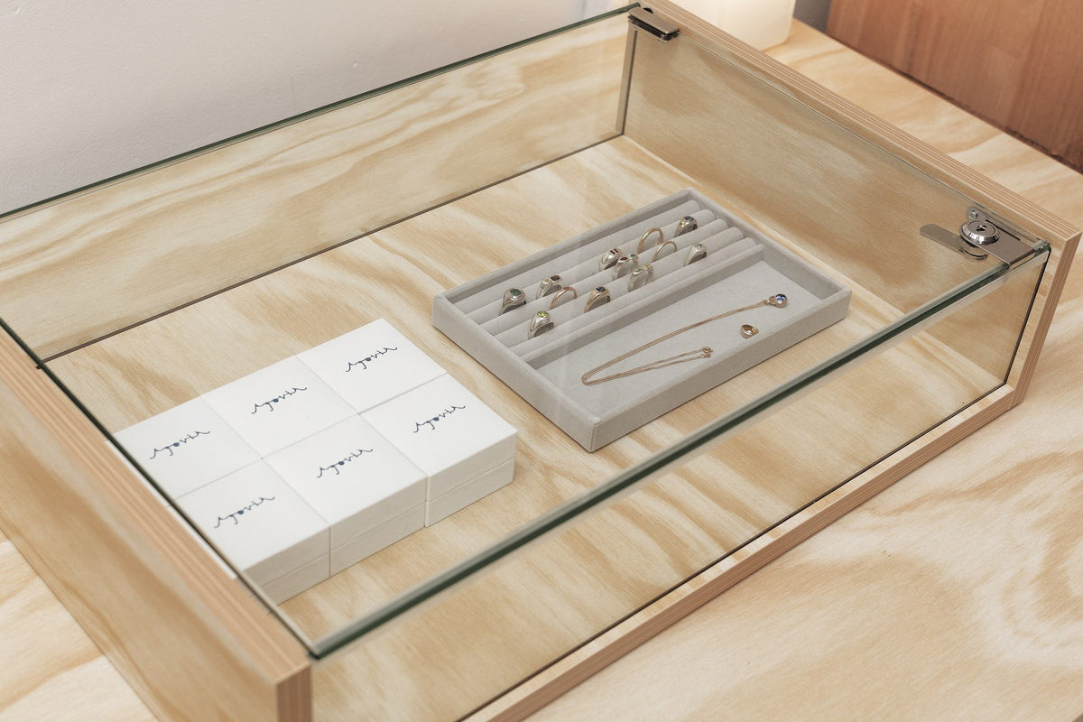Rings and a fine gold chain on a tray inside the plywood and glass display case, beside stacked AGOVIA jewellery boxes.